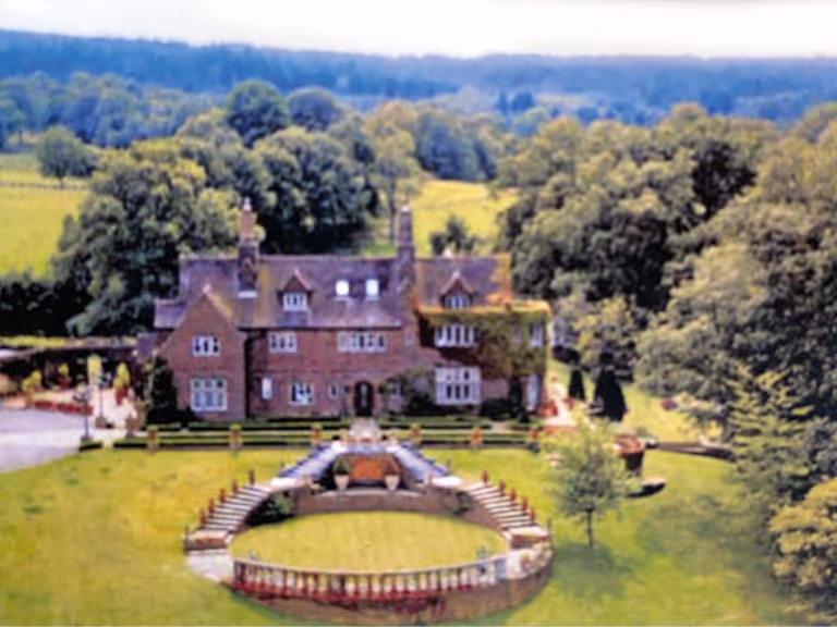 Large brick house surrounded by lush greenery and trees with a circular stone-bordered garden in front.