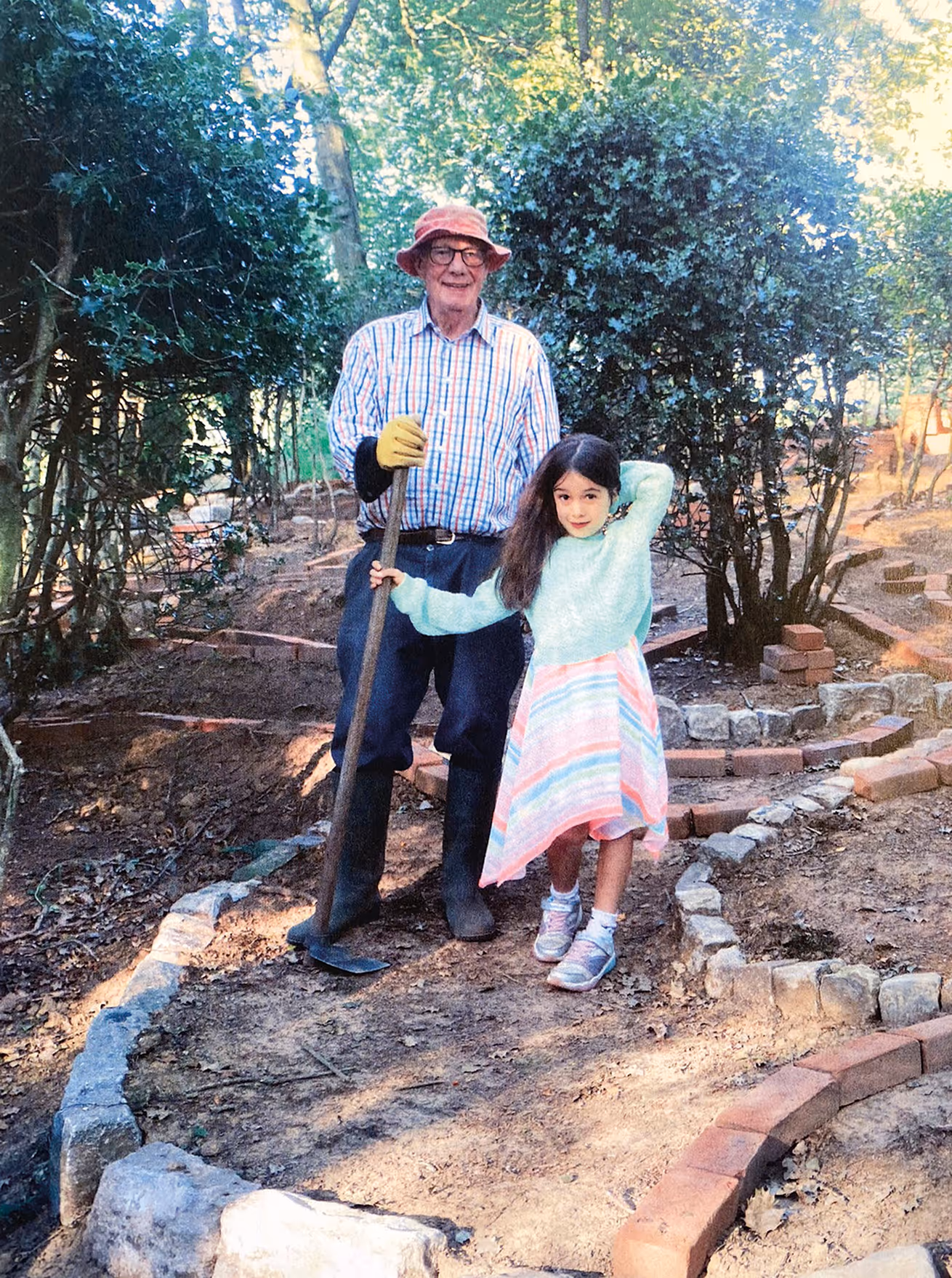Philip Wood wearing a hat and gloves stands with a gardening hoe next to a young girl in a blue sweater and striped skirt in a garden with newly laid brick borders.