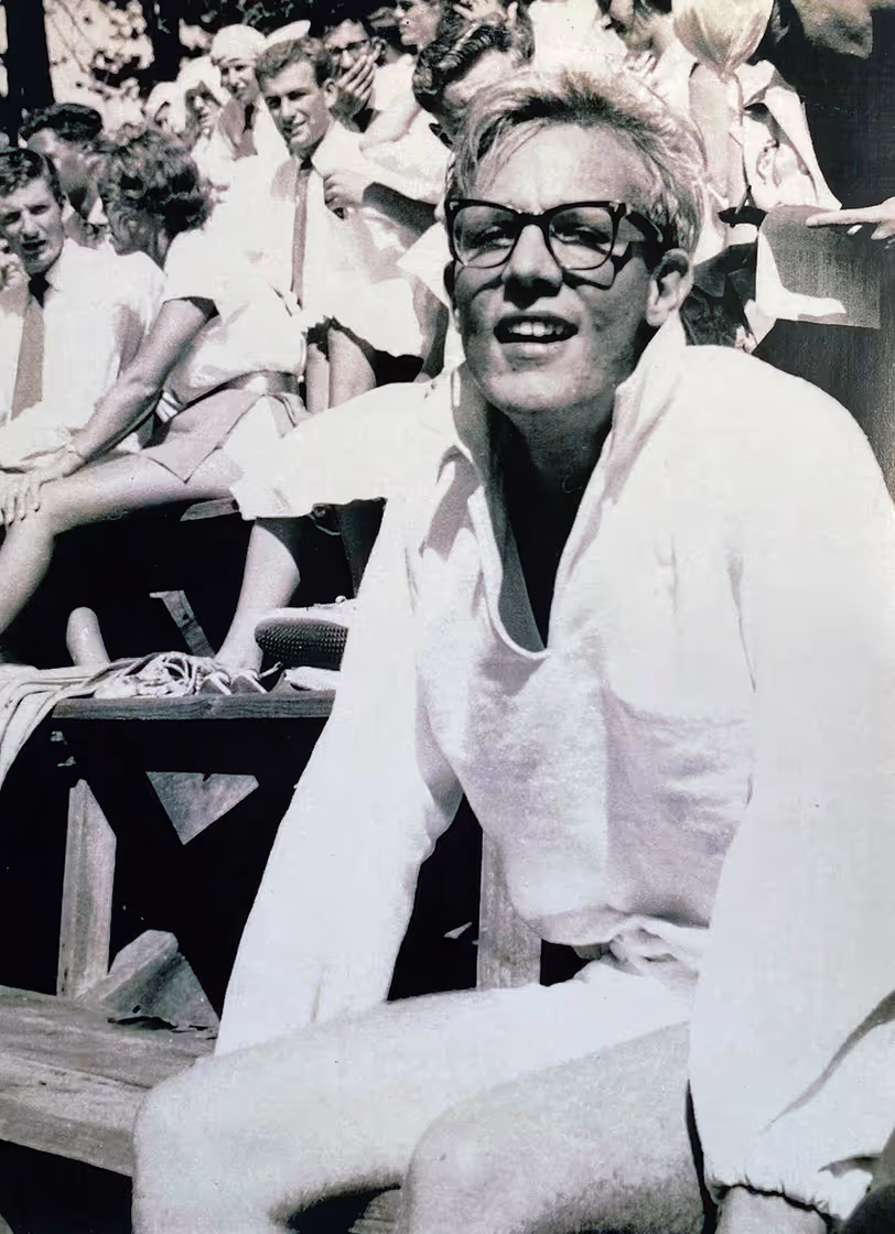 Black and white photo of a young Philip Wood wearing glasses and light-colored clothing sitting on wooden bleachers with other people in the background.