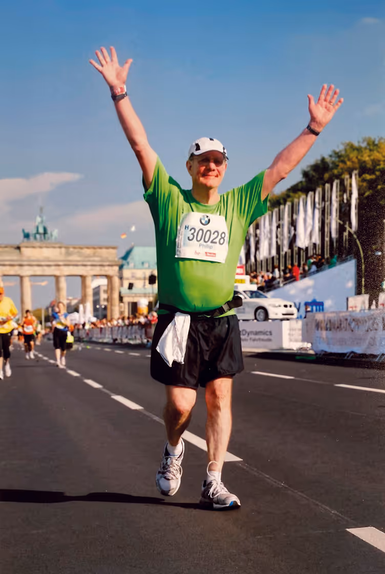 Philip Wood in green shirt and black shorts raising both arms while running near Brandenburg Gate in Berlin.