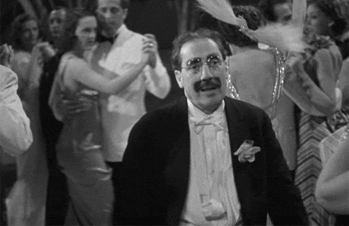 Black and white scene of a man with glasses and a mustache dancing amid couples ballroom dancing in formal attire.