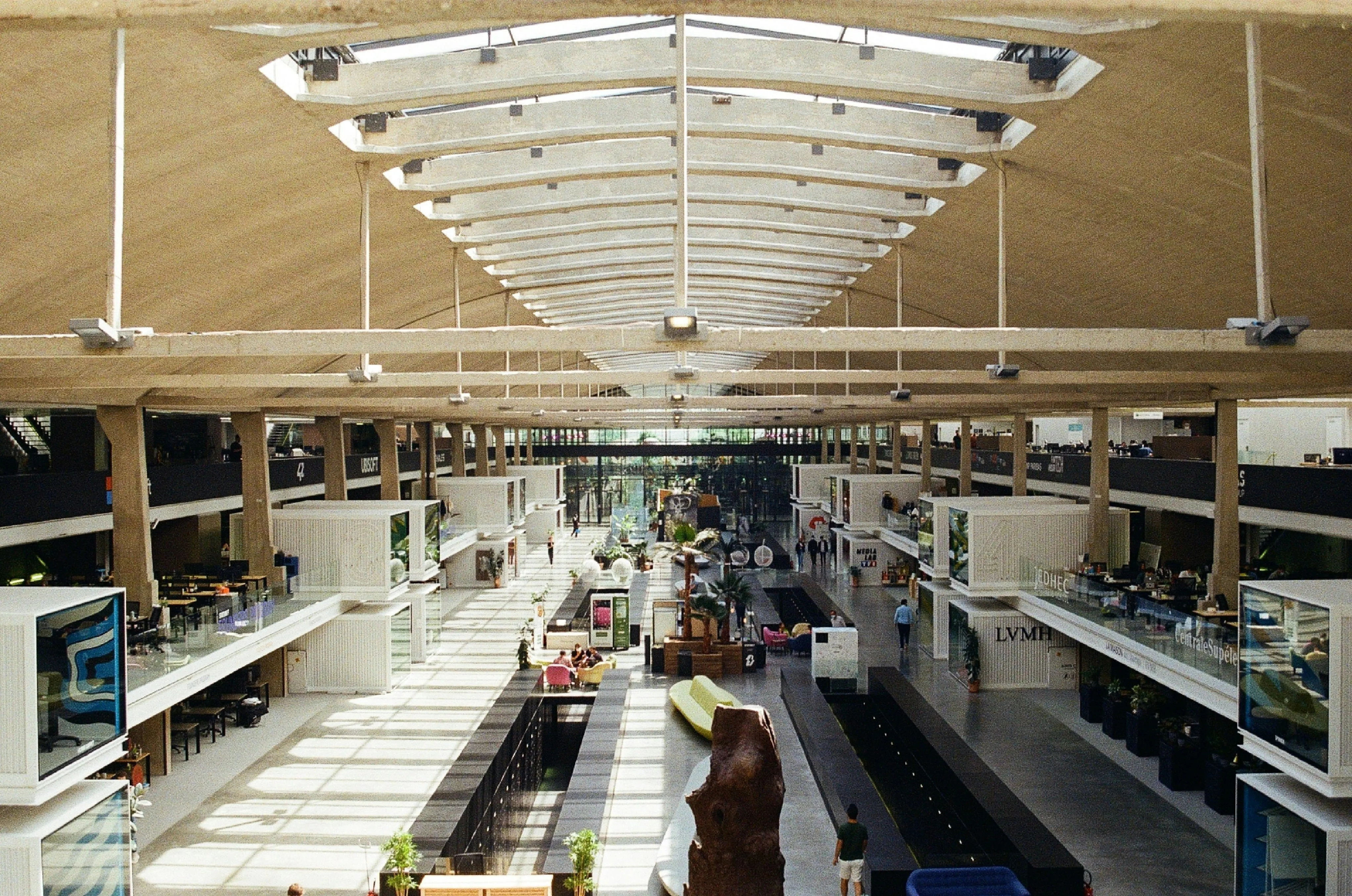 Spacious modern interior of a building with high ceiling, skylights, walkways, seating areas, and people walking inside.