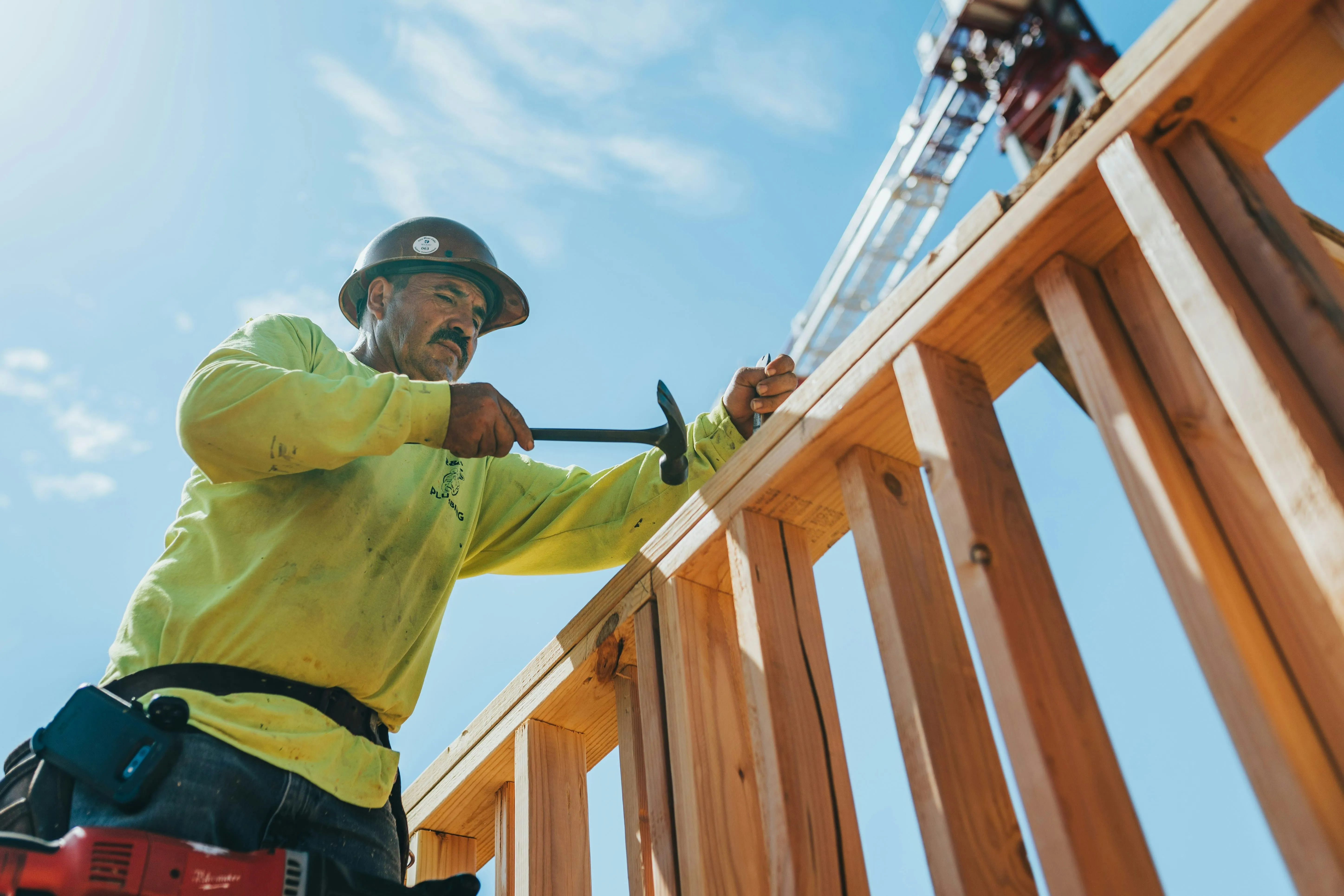 Construction worker wearing a hard hat and yellow shirt hammering a nail into wooden framing on a bright sunny day.
