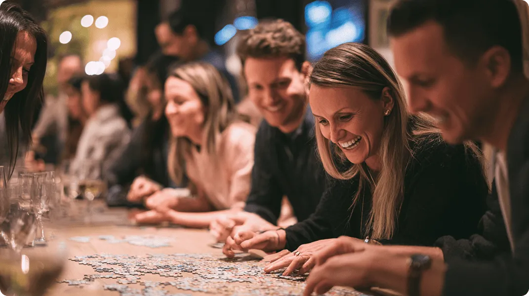 Group of people happily assembling a jigsaw puzzle on a table in a cozy indoor setting.