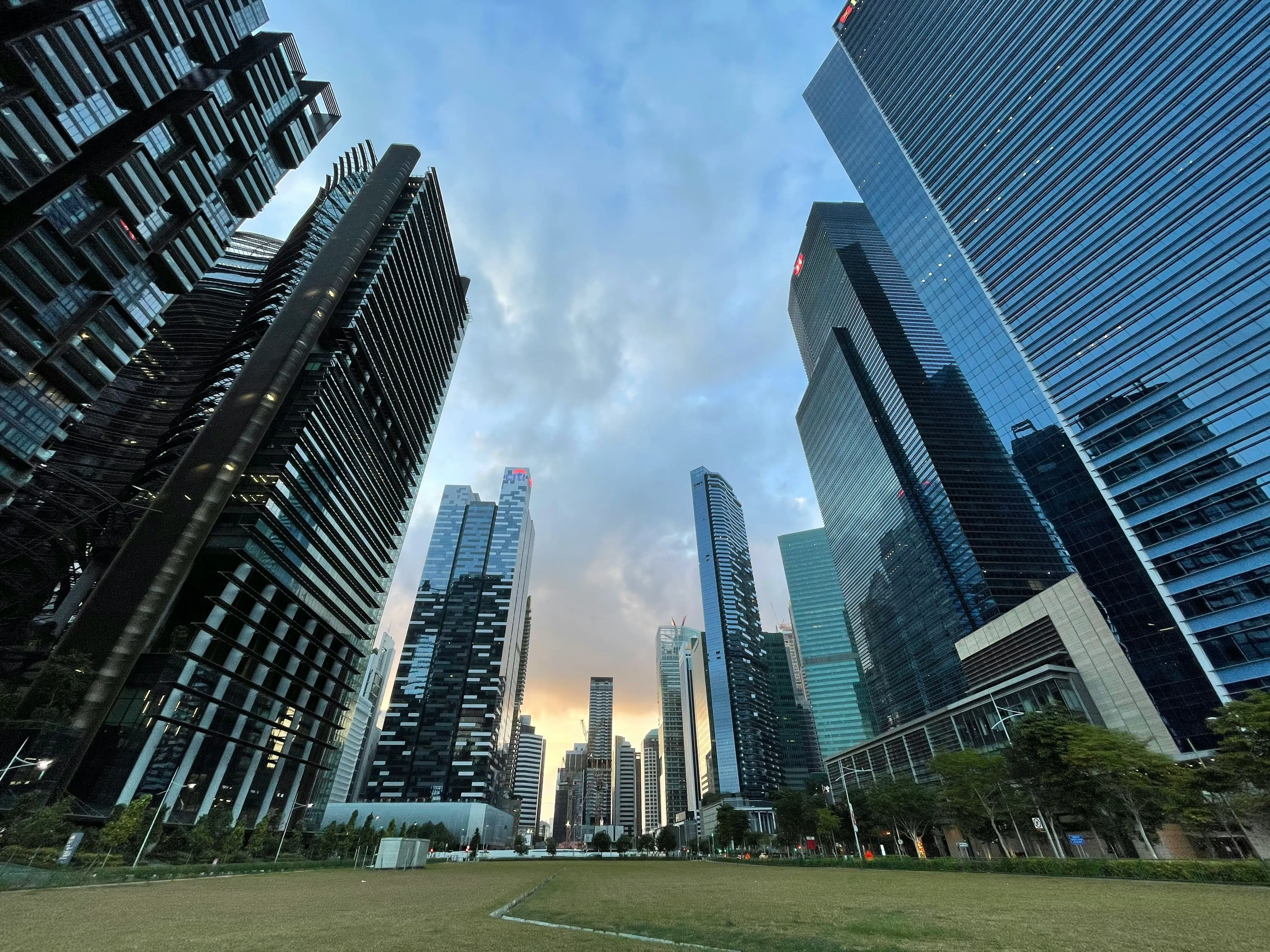 View of modern skyscrapers surrounding a green lawn at sunset under a partly cloudy sky.