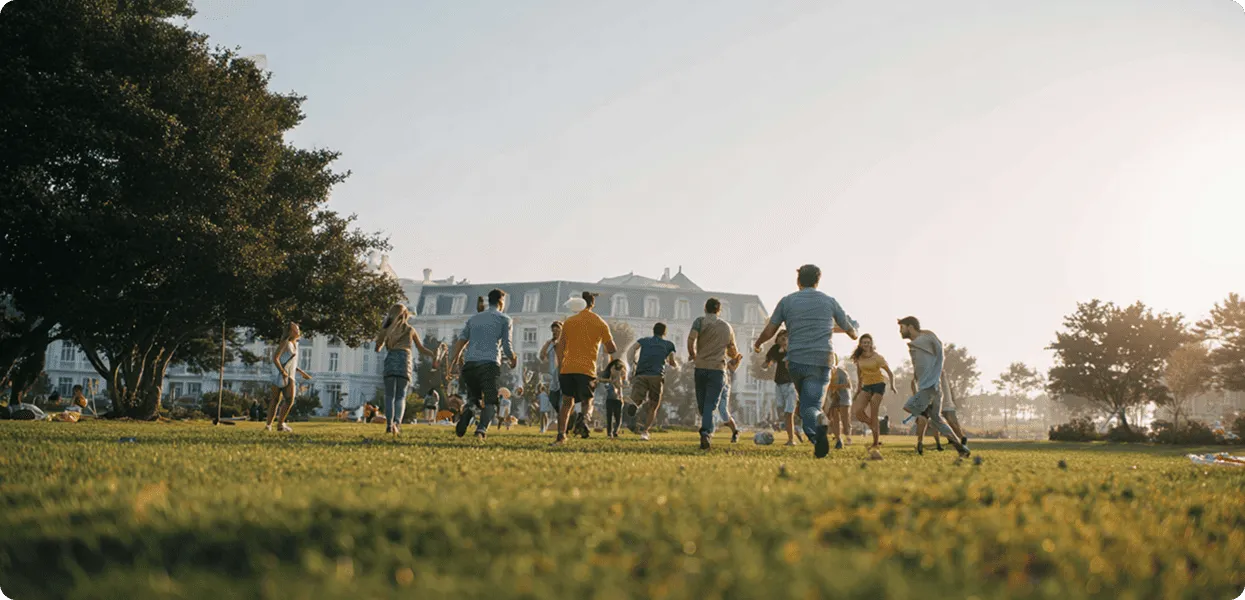 Group of people playing soccer on a sunny grassy field near large buildings and trees.