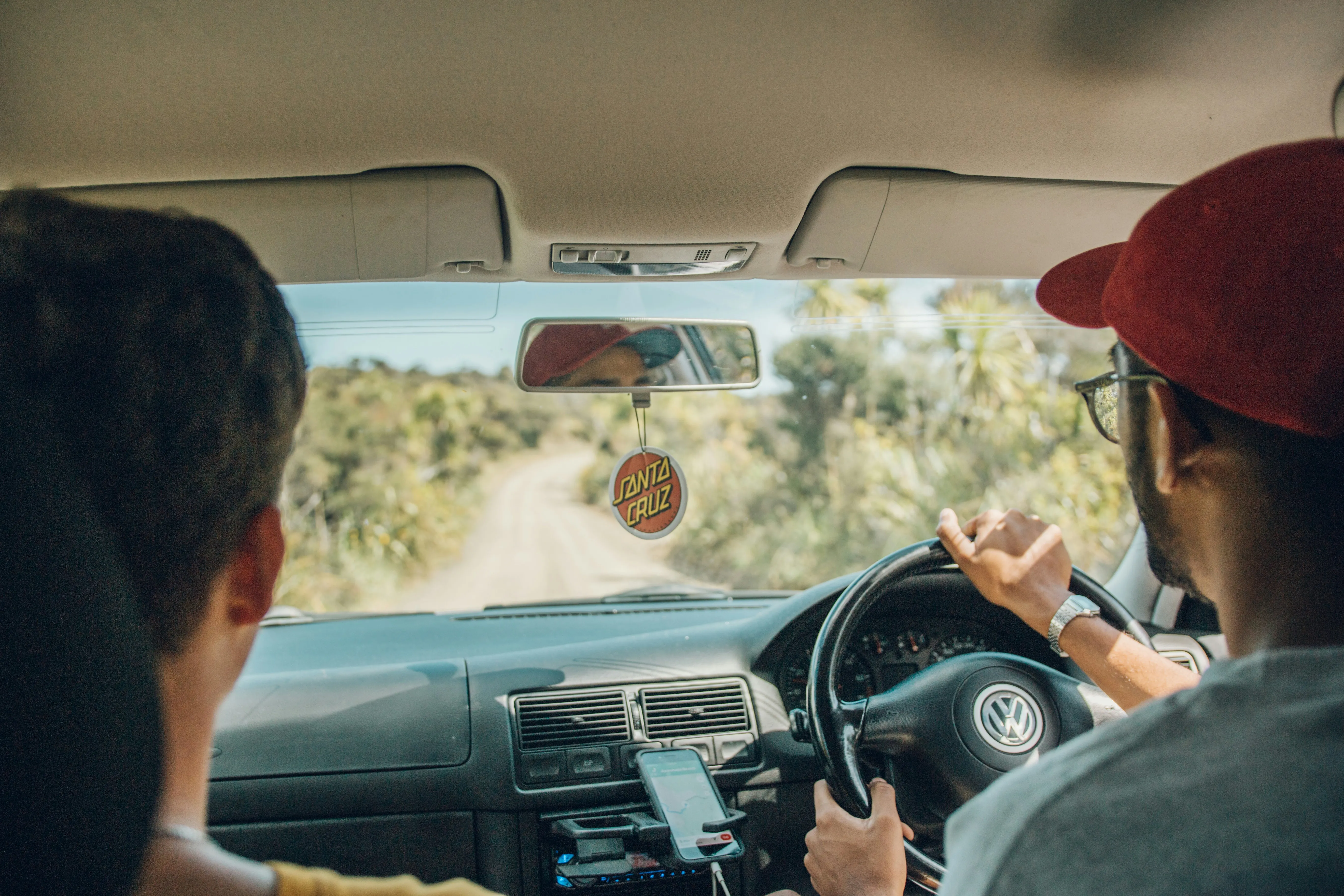 Two men inside a Volkswagen car driving on a dirt road surrounded by greenery, with a Santa Cruz air freshener hanging from the rearview mirror.