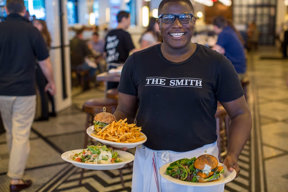 Smiling waiter wearing glasses and a black 'THE SMITH' t-shirt carrying plates with burgers, fries, and salad in a busy restaurant.