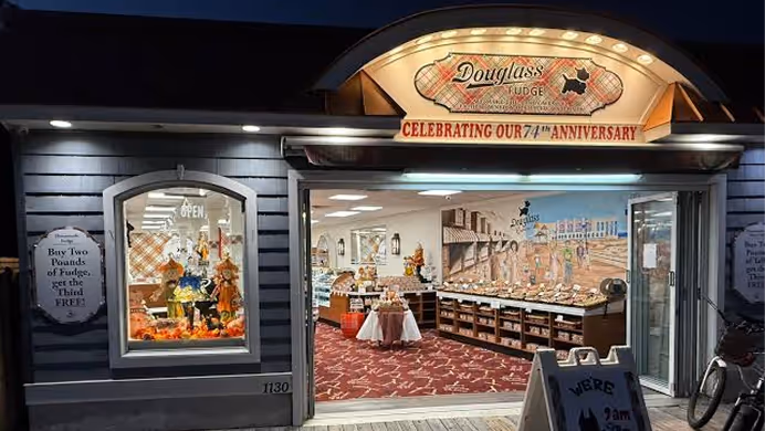 Exterior of Douglass Fudge store at night with open entrance, displaying fudge and decorations inside.