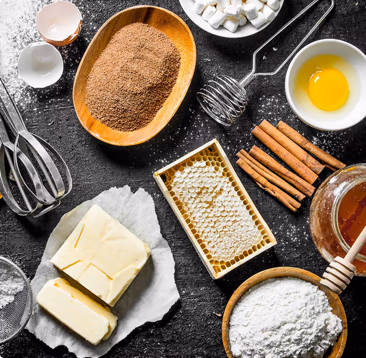 Various baking ingredients and tools on a dark surface, including butter, powdered sugar, cinnamon sticks, an egg yolk, marshmallows, honeycomb, cocoa powder in a wooden bowl, egg shells, and metal whisks.