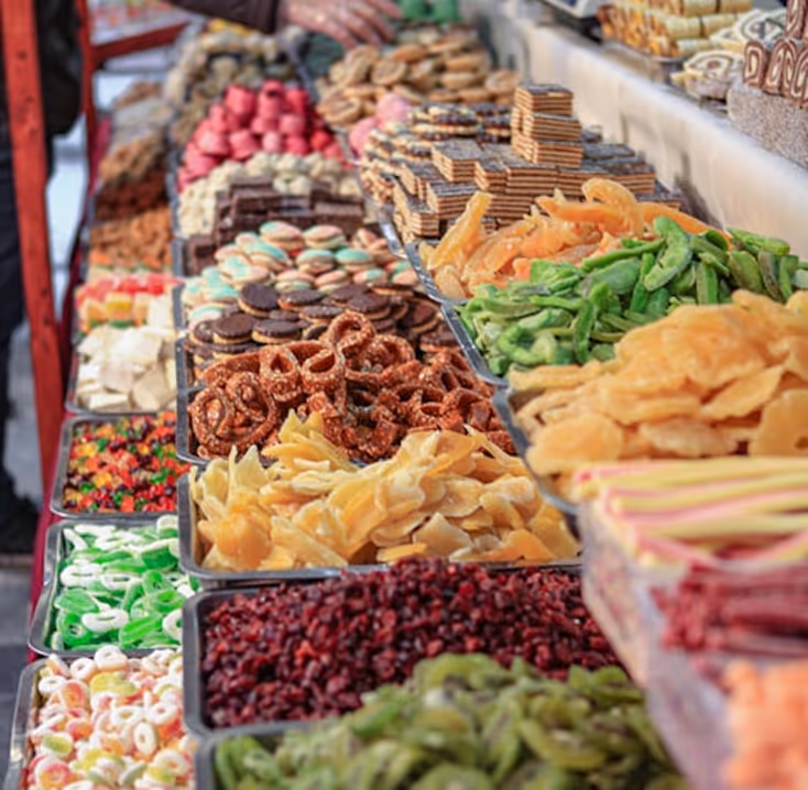 Market display with trays full of assorted colorful candies, dried fruits, pretzels, and cookies.