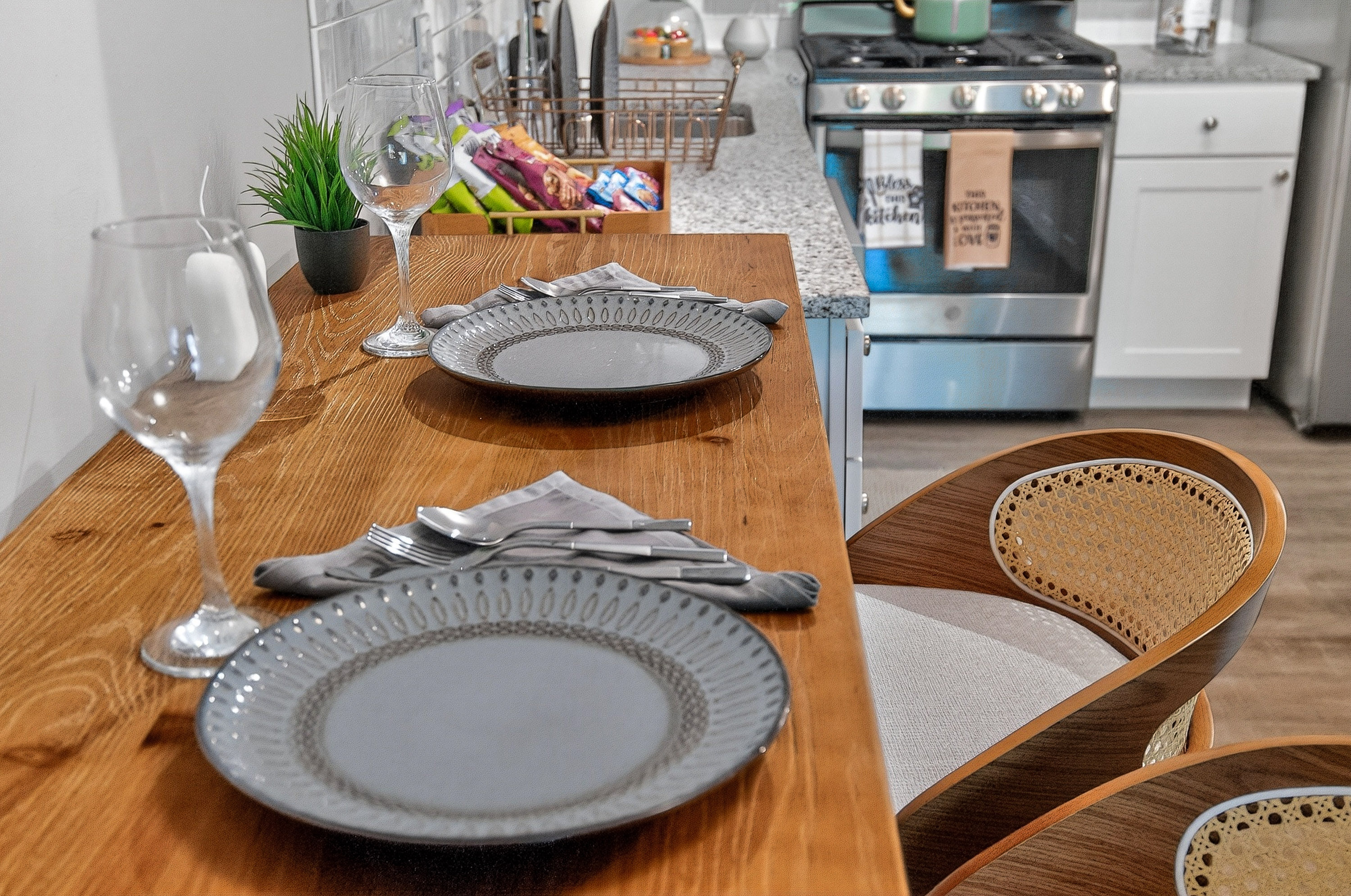 Wooden kitchen counter set with two plates, wine glasses, silverware, and curved chairs with woven backs.