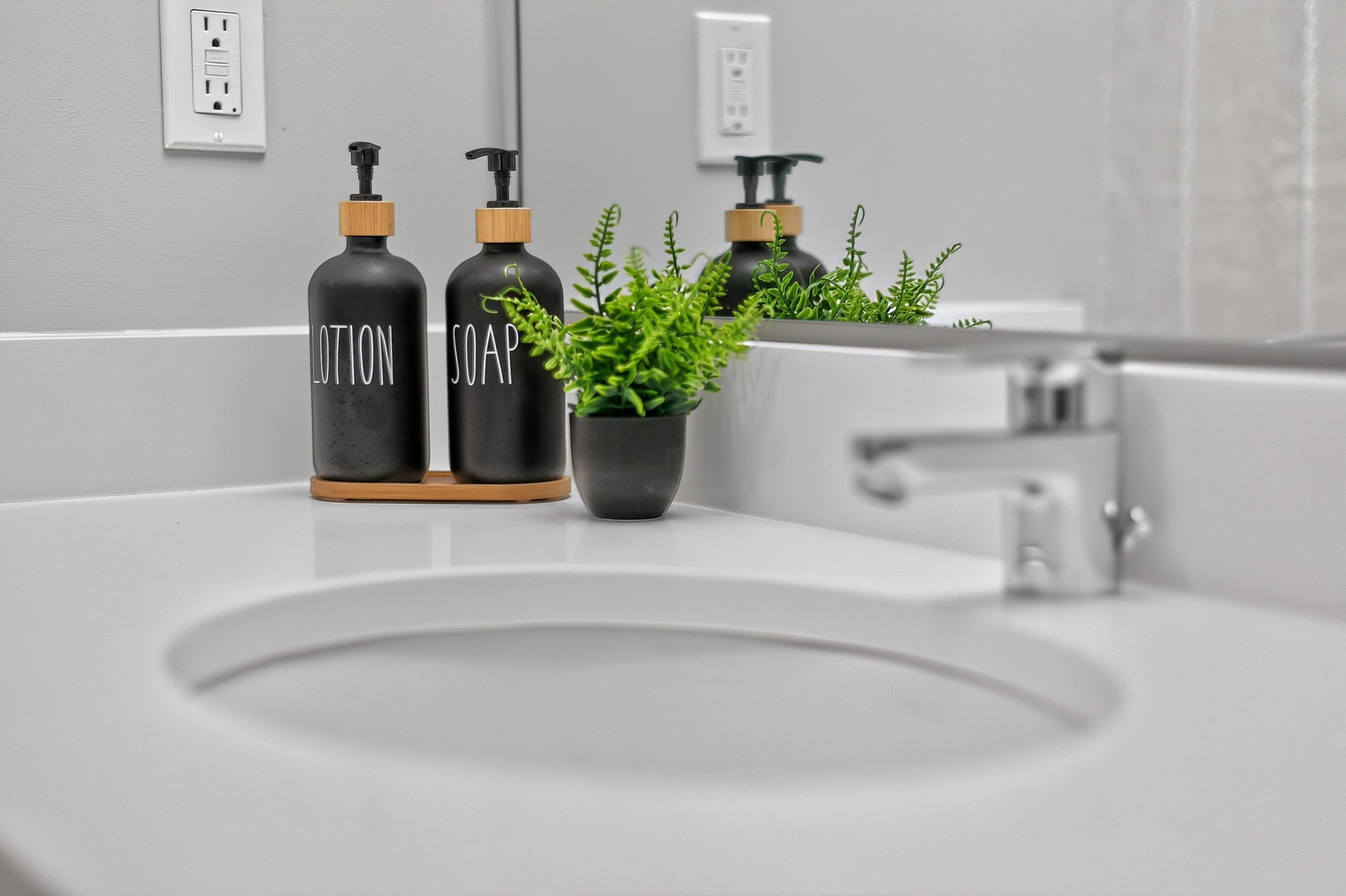 White bathroom sink with chrome faucet, black lotion and soap dispensers, and small green plant on counter.