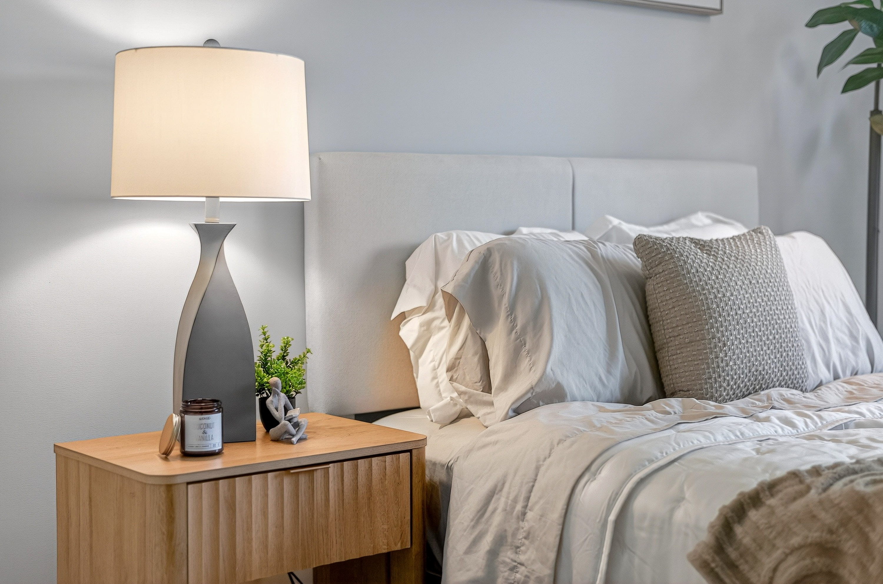 Nightstand with lit lamp, candle, small plant, and gray figurine next to bed with white bedding.