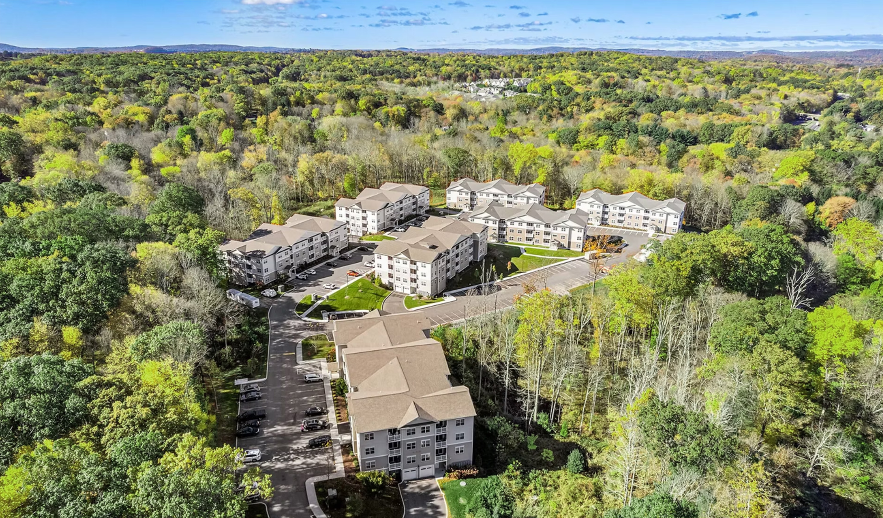 Aerial view of a residential apartment complex surrounded by dense green forest under blue sky.