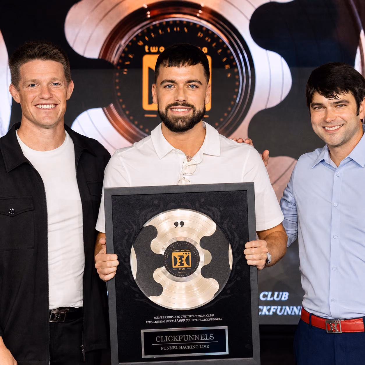 Three men smiling, with the man in the center holding a ClickFunnels Two-Comma Club award plaque.