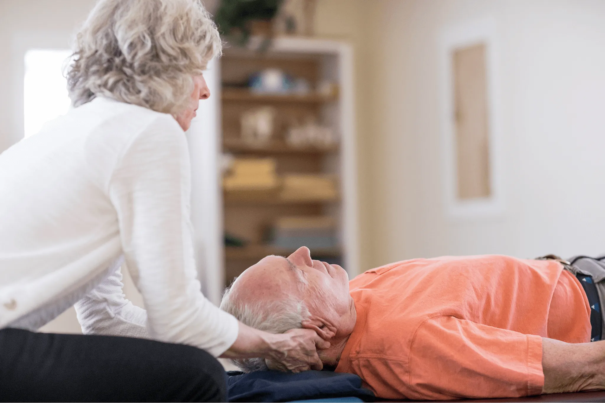 Lindy Ost supporting the neck of an elderly man lying down in a calm indoor setting.