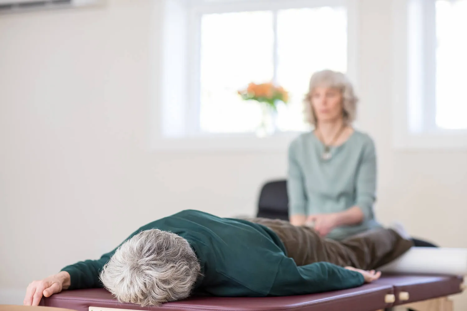 Senior person lying face down on a therapy table with a practitioner sitting nearby in a bright room.