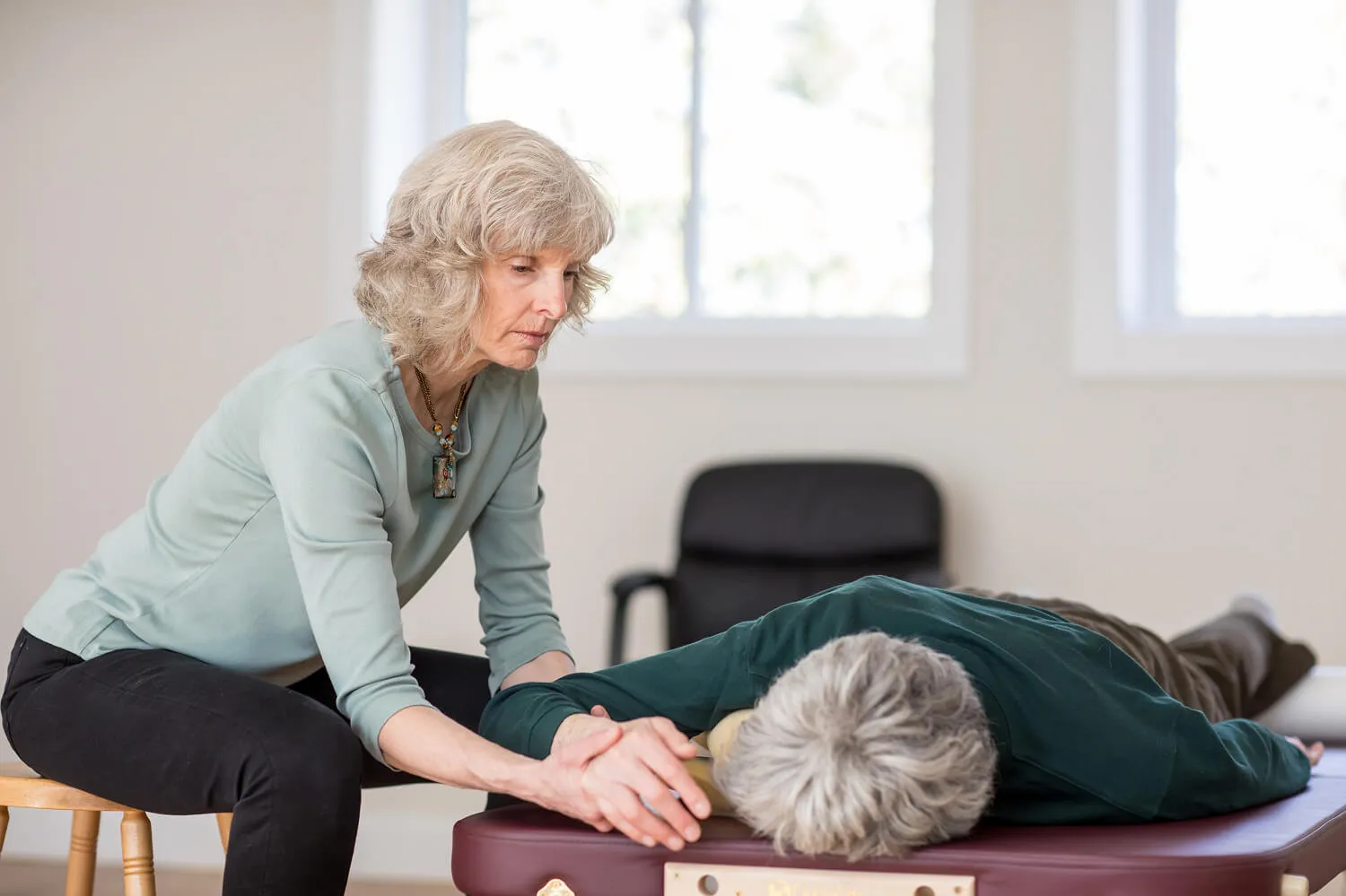 Woman with gray hair assisting an elderly person lying face down on a therapy table in a bright room.