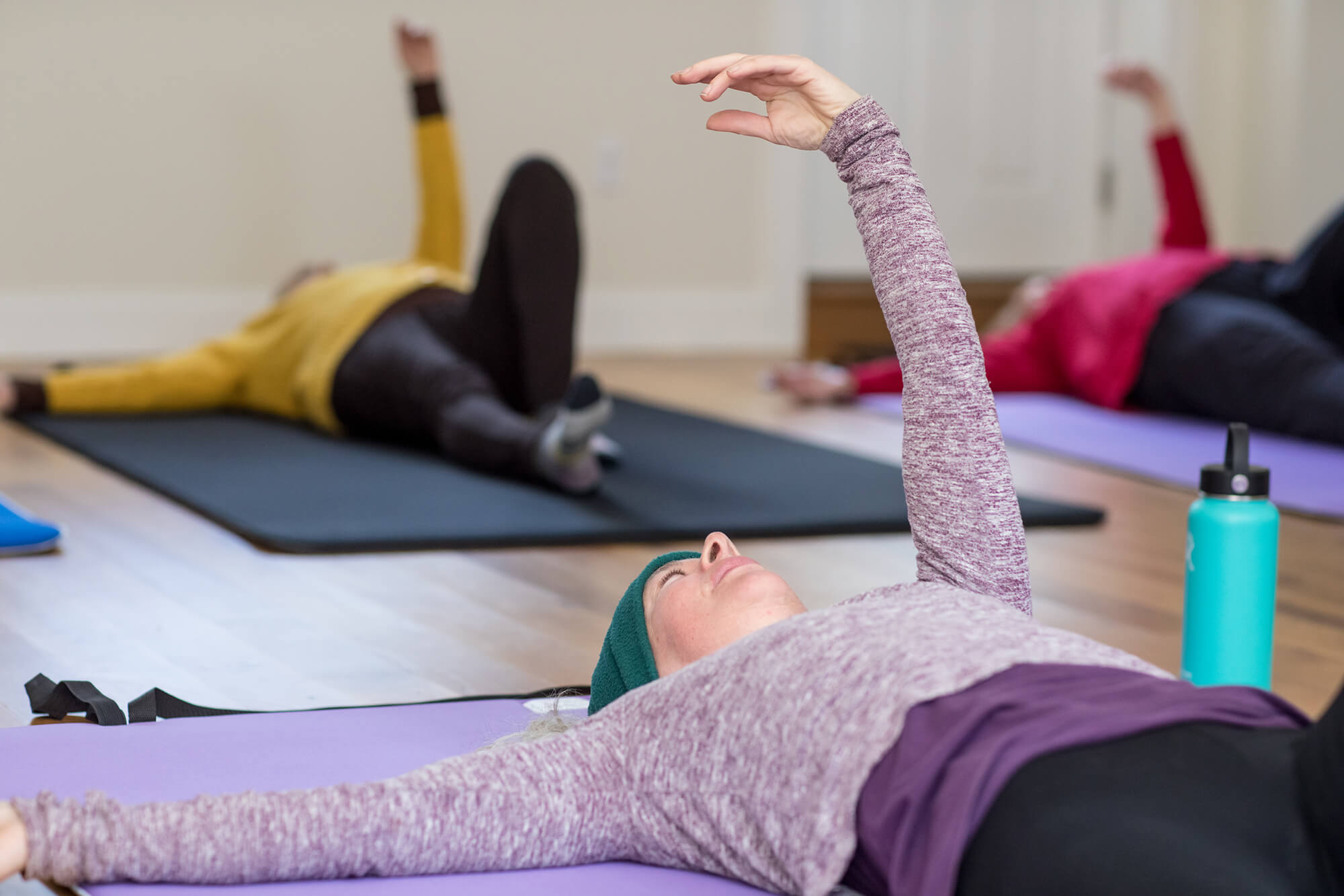 Three people lying on exercise mats indoors, stretching one arm upward during a group fitness class.