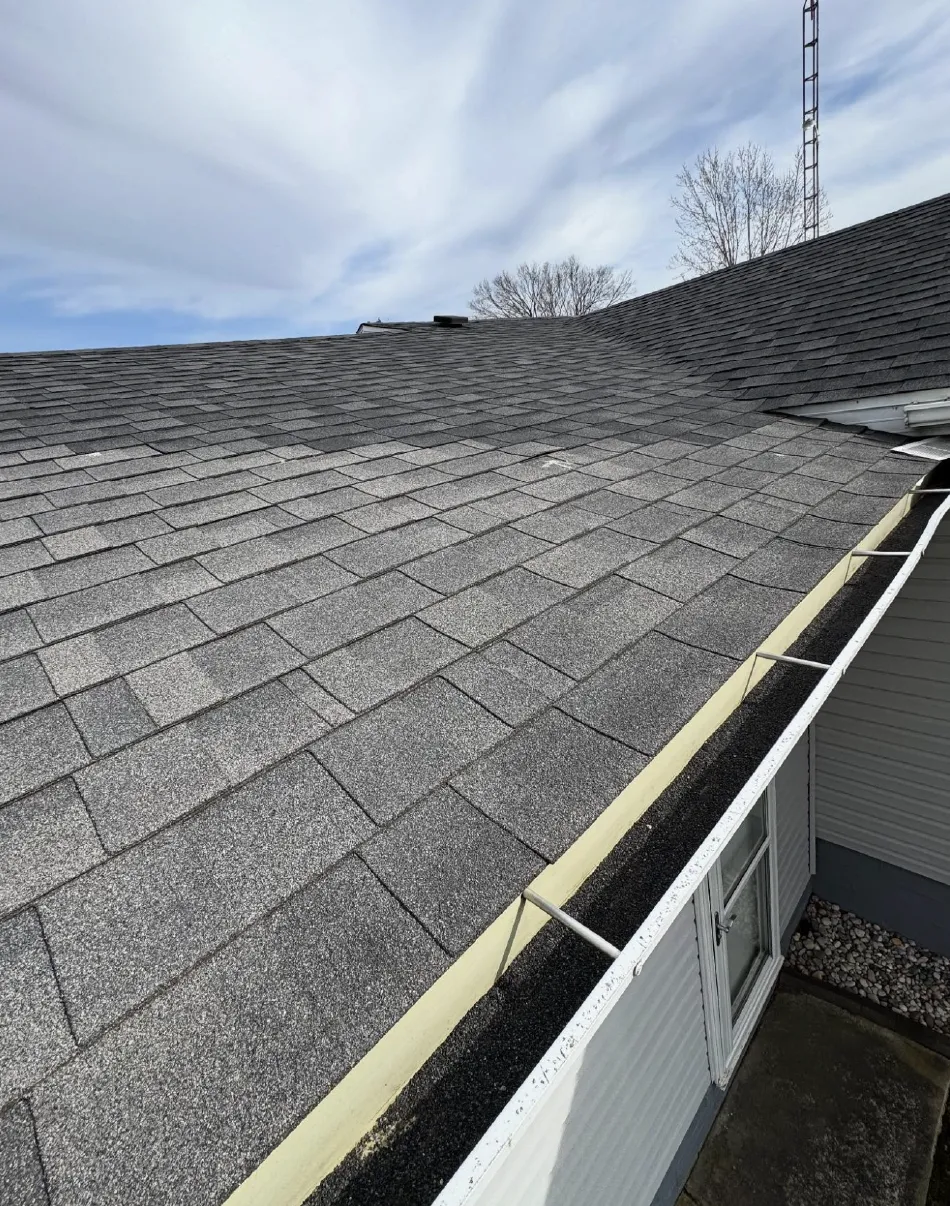 View of a gray shingled roof with a clean white gutter attached to a white house exterior under a cloudy sky.