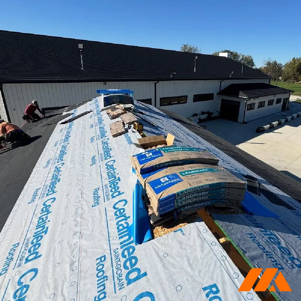 Two workers installing roofing underlayment and shingles on a commercial building roof under a clear blue sky.