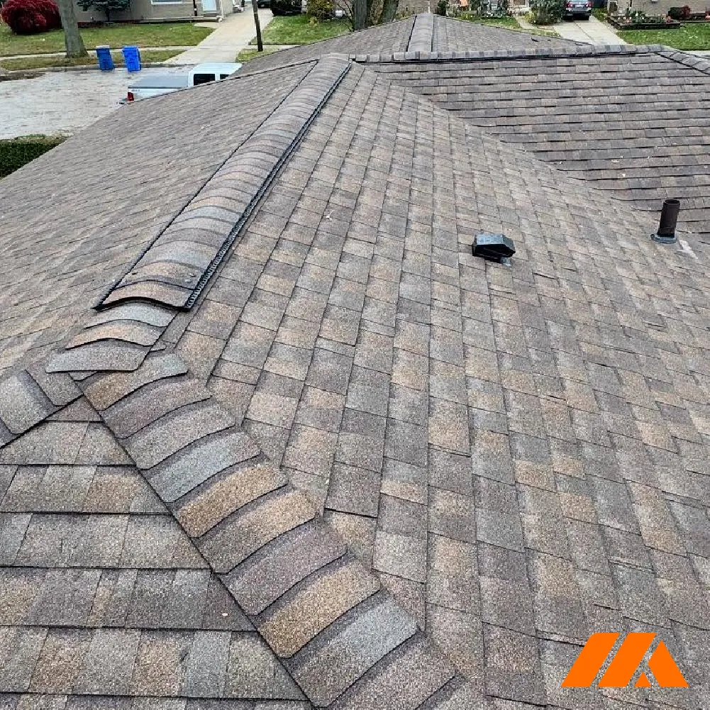 View of a residential roof with gray and brown asphalt shingles and ridge caps on a cloudy day.
