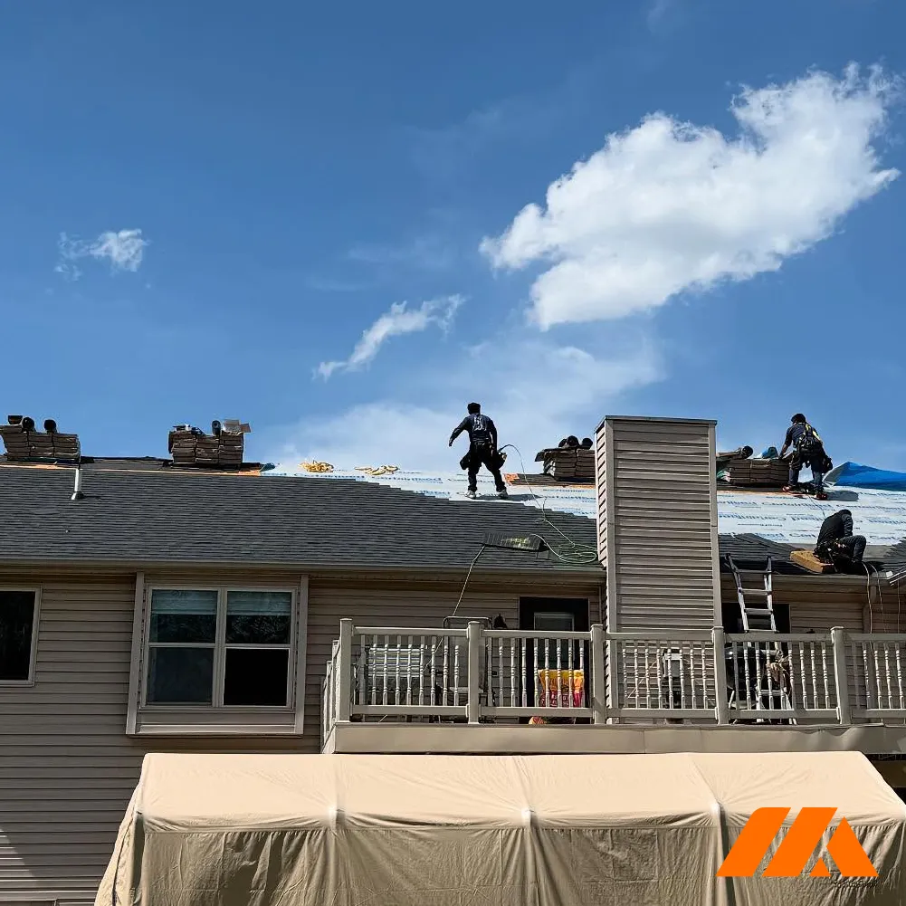 Workers installing new roofing shingles on a residential house under a blue sky with white clouds.