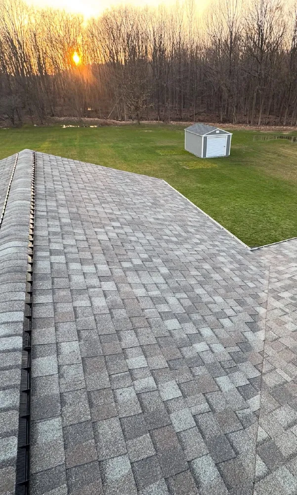 Gray shingled roof of a house overlooking a green yard with a small gray shed, with bare trees and a sunset in the background.
