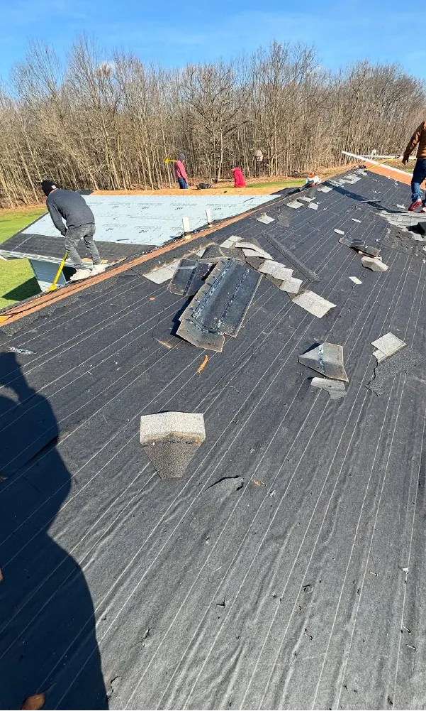 Roof under construction with black roofing underlayment, scattered shingles, and workers installing roofing materials under clear blue sky.