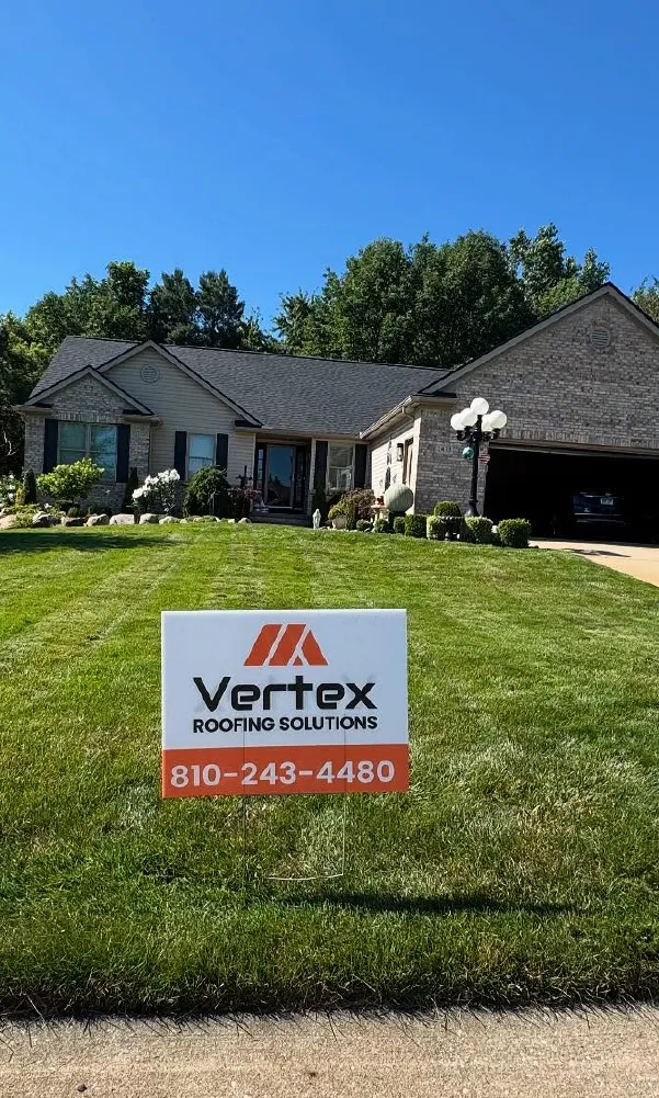 Lawn with a Vertex Roofing Solutions sign featuring phone number 810-243-4480 in front of a suburban house with brick and siding exterior under a clear blue sky.