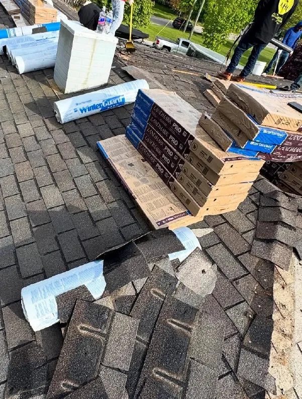 Stacks of roofing shingles and rolls of underlayment on a roof with workers preparing for installation.