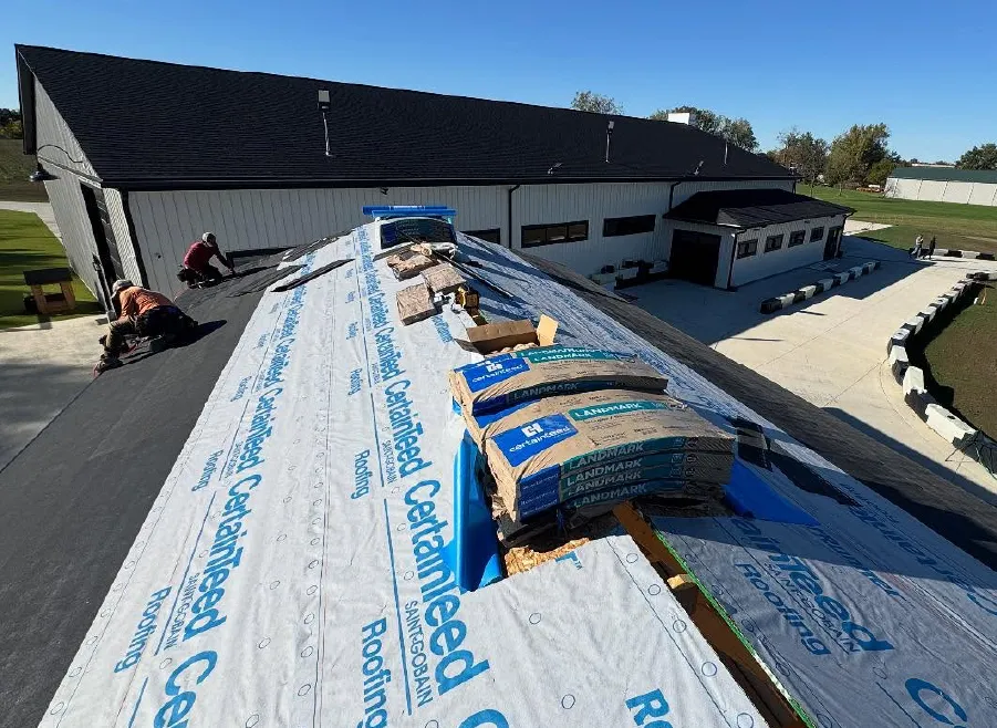 Workers installing roofing materials on a large building under clear blue sky, with stacks of shingles and protective underlayment visible.