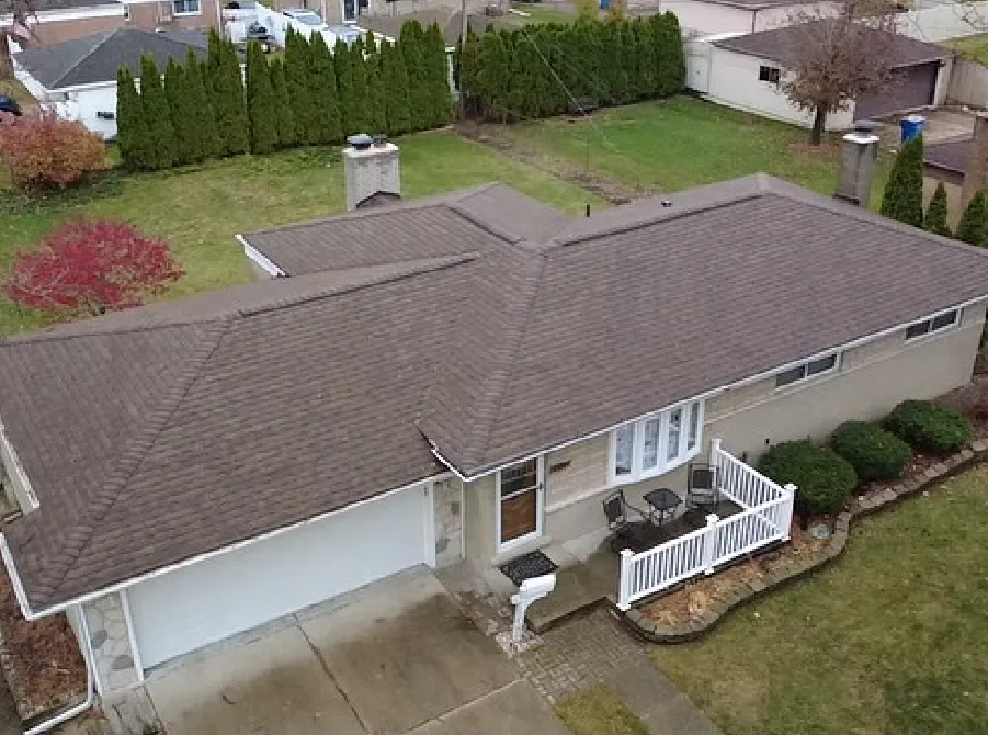Aerial view of a single-story house with a brown shingle roof, attached garage, small front porch with white railing, and surrounding green lawn with shrubs and trees.