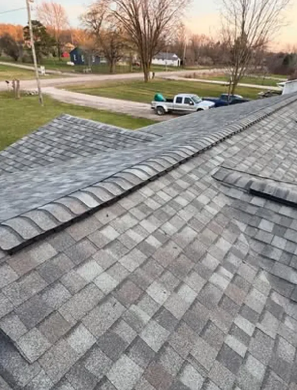 A gray shingled residential roof with a raised ridge and a view of a suburban street with parked pickup trucks and bare trees.