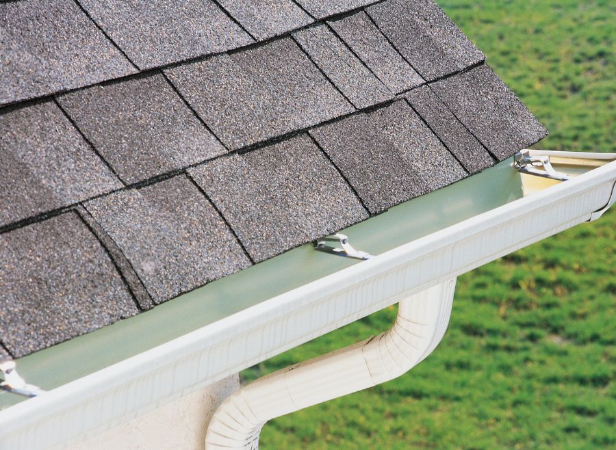 Close-up of house roof with asphalt shingles and attached white rain gutter and downspout on a sunny day.