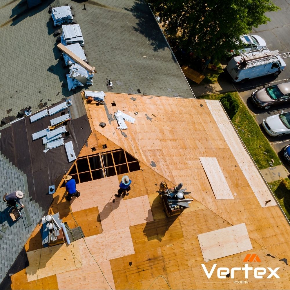Aerial view of workers installing plywood roofing panels on a house under construction with roofing materials stacked nearby.