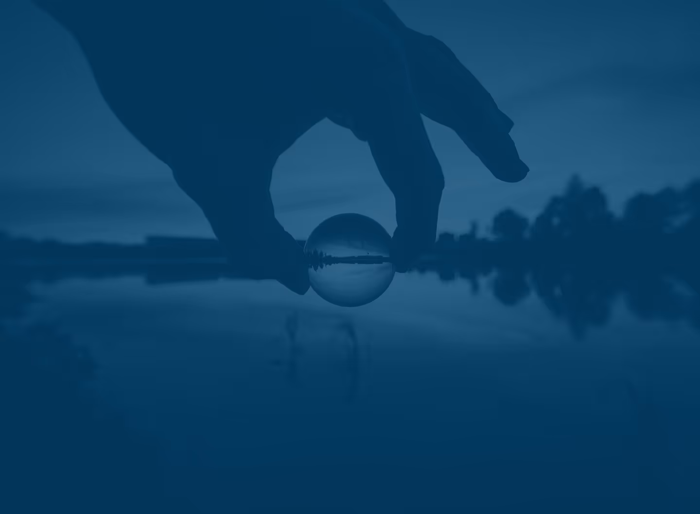 Hand holding a small glass sphere reflecting an inverted image of a lake and trees at dusk.