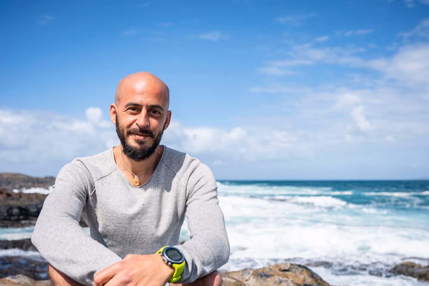 Smiling bald man with a beard wearing a gray sweater and yellow watch sitting on rocks by the ocean under a blue sky.