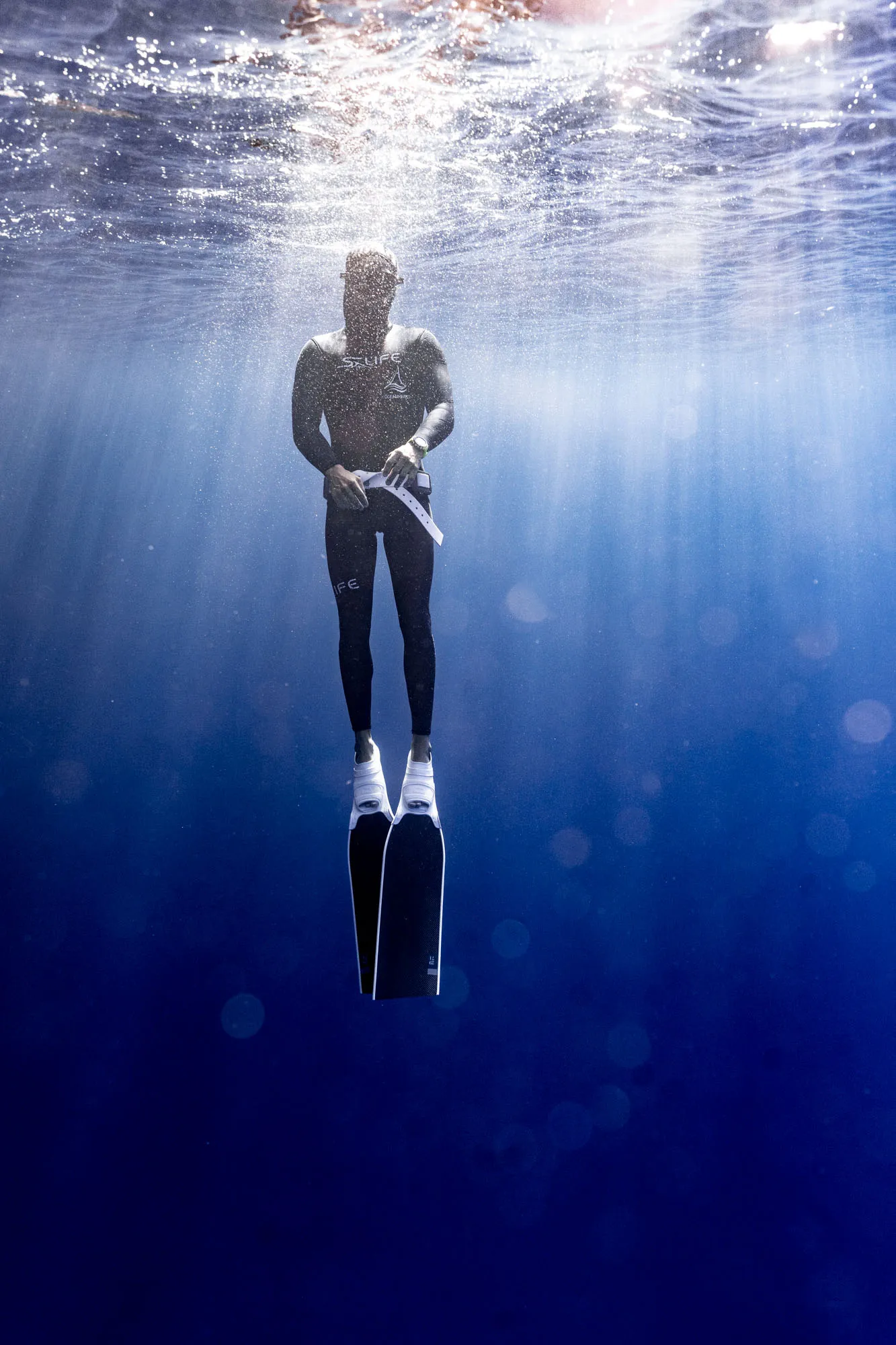 Person in a wetsuit with diving fins floating underwater with sunlight filtering from above.