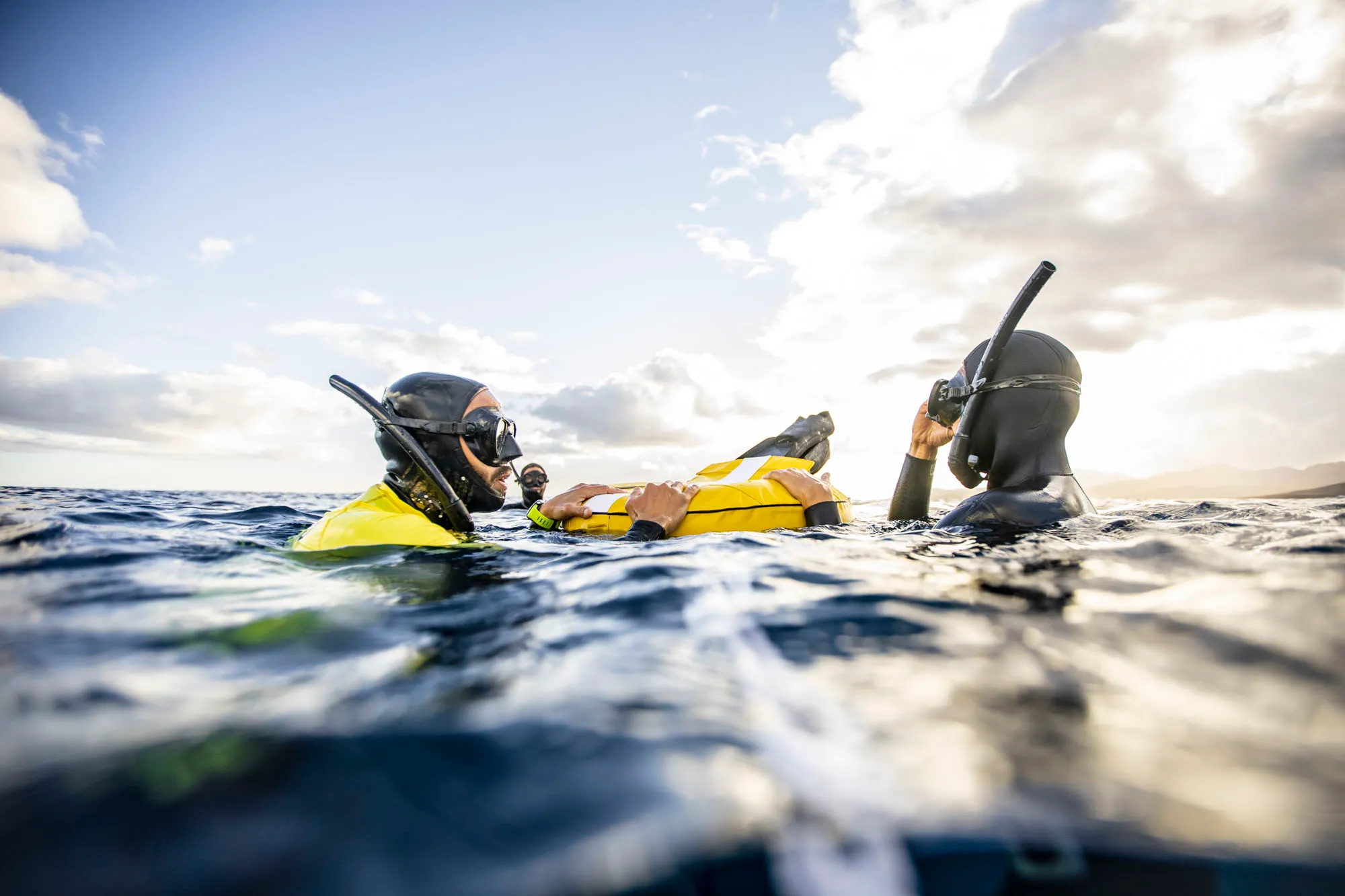 Three snorkelers in wetsuits floating on the ocean surface holding a yellow flotation device under a partly cloudy sky.
