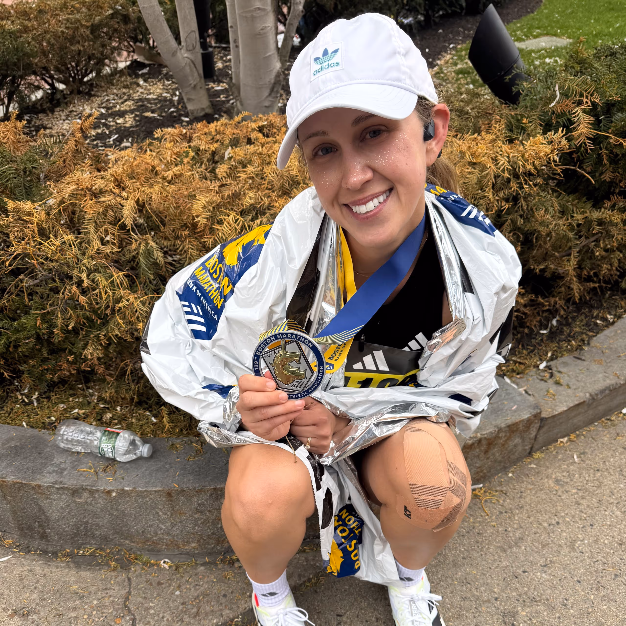 Smiling female runner wearing a white Adidas cap and knee tape holds a Boston Marathon finisher's medal wrapped in a foil blanket.