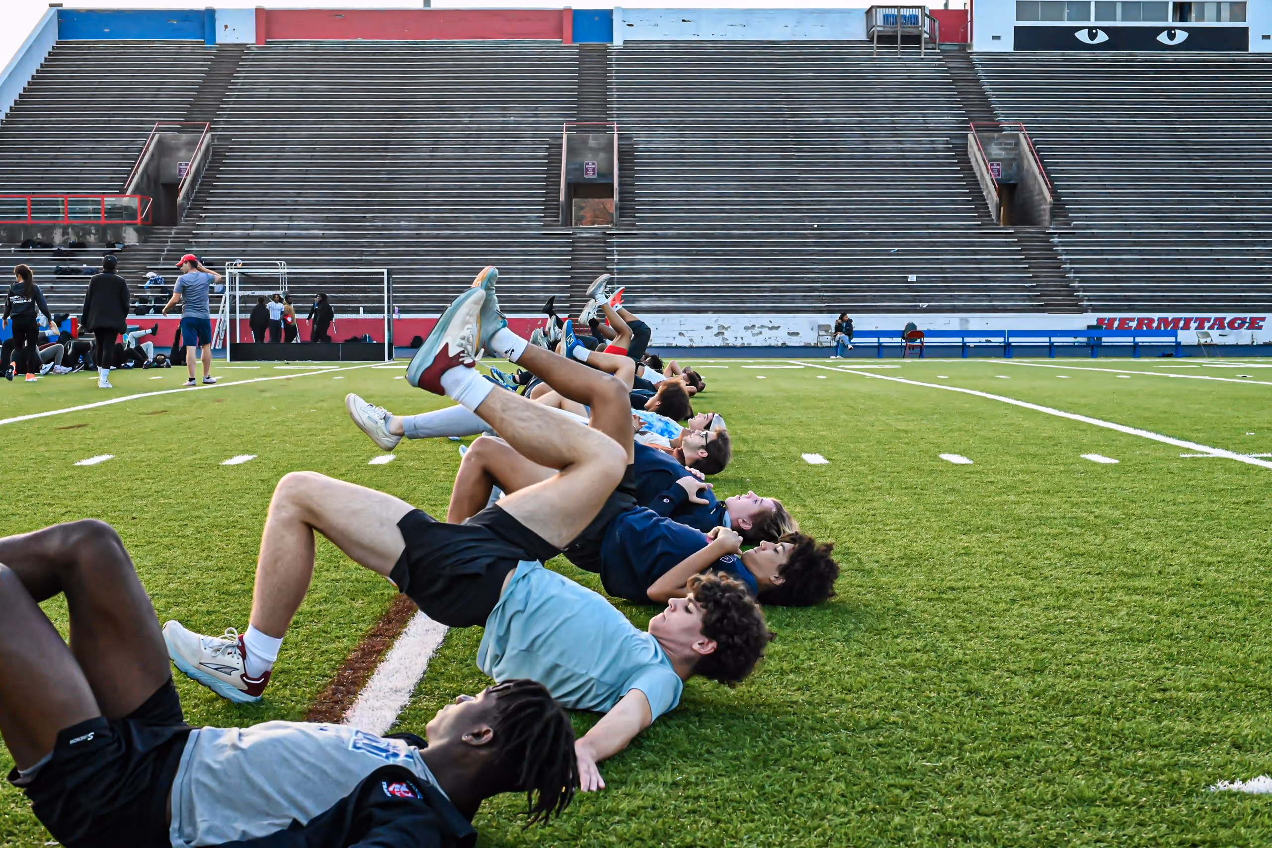 Group of young athletes lying on their backs on a football field, lifting one leg during a warm-up exercise.