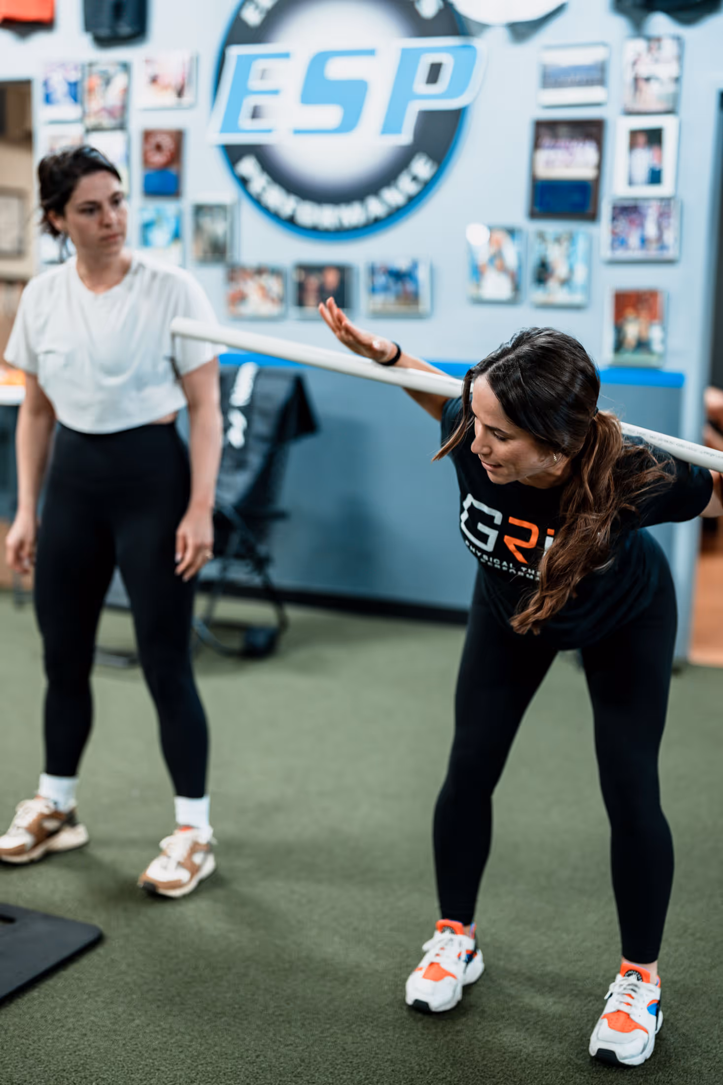 A physical therapist in exercise attire bends forward holding a barbell across her shoulders while the patient watches her in a richmond physical therapy clinic.