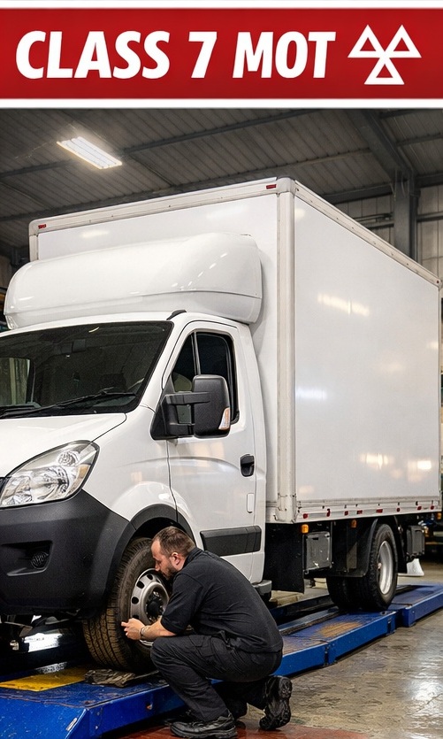 Mechanic inspecting the front tyre of a white commercial van k on a vehicle lift under a red banner reading 'CLASS 7 MOT'.