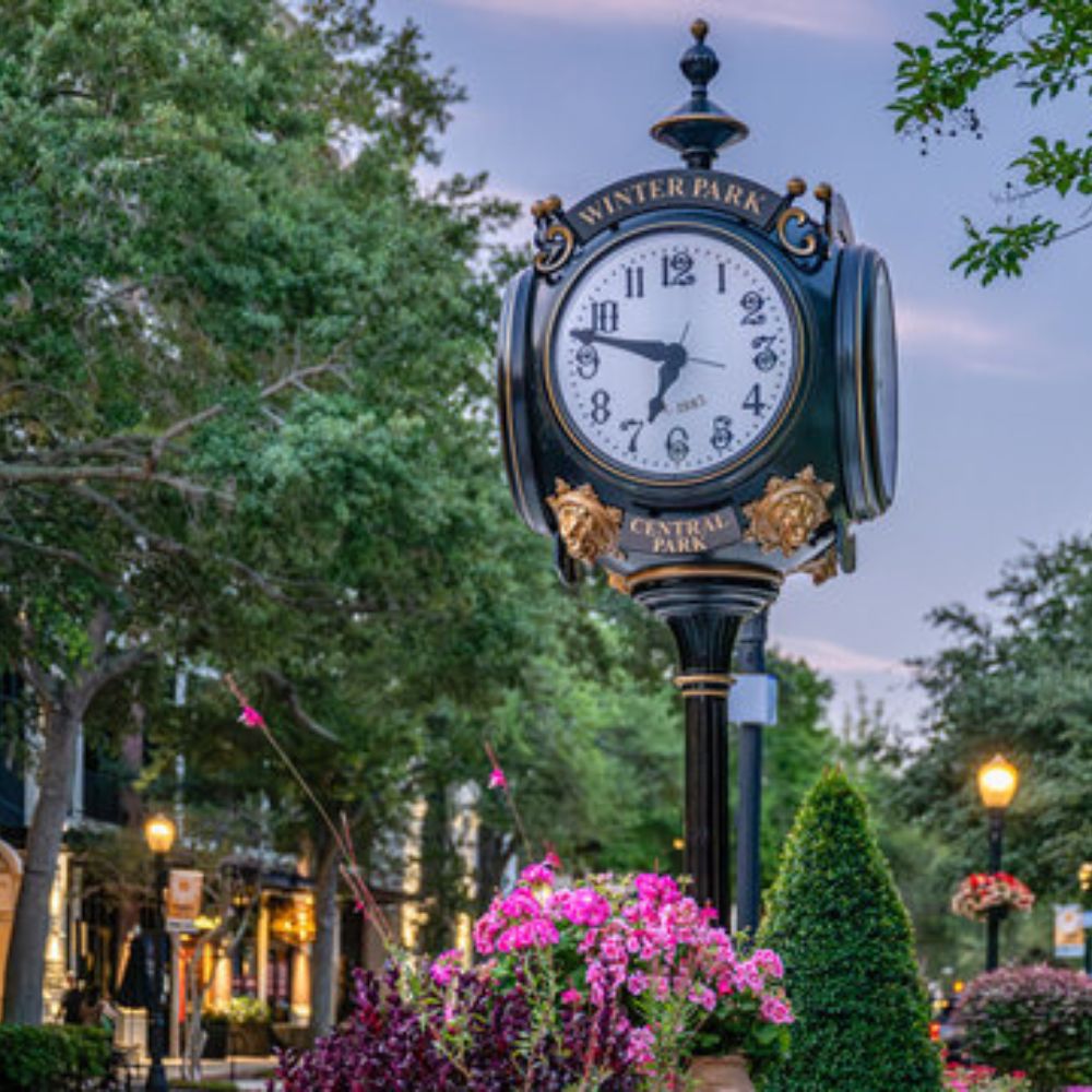 Winter Park clock on Central Park at dusk, with pink flowers and street lamps along the sidewalk