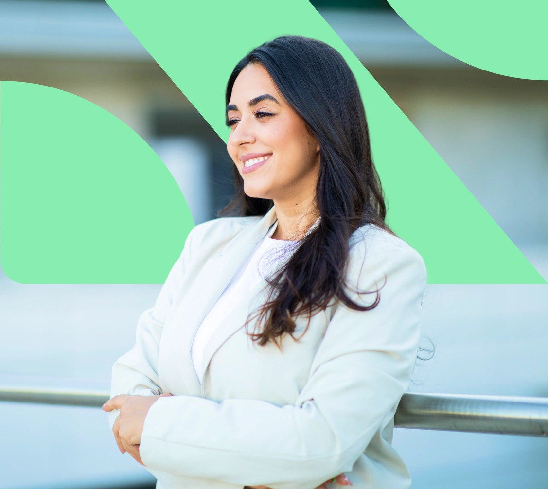 Smiling woman with long dark hair wearing a white blazer, standing with arms crossed outdoors.