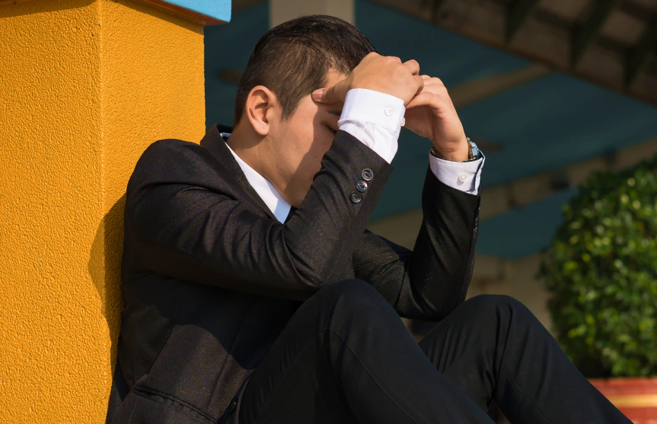 Man in a black suit sitting against a yellow wall with his head in his hands, appearing stressed or overwhelmed.
