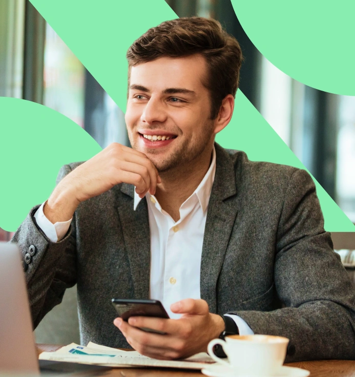 Smiling man in a gray blazer holding a smartphone, sitting at a table with a laptop and a cup of coffee.