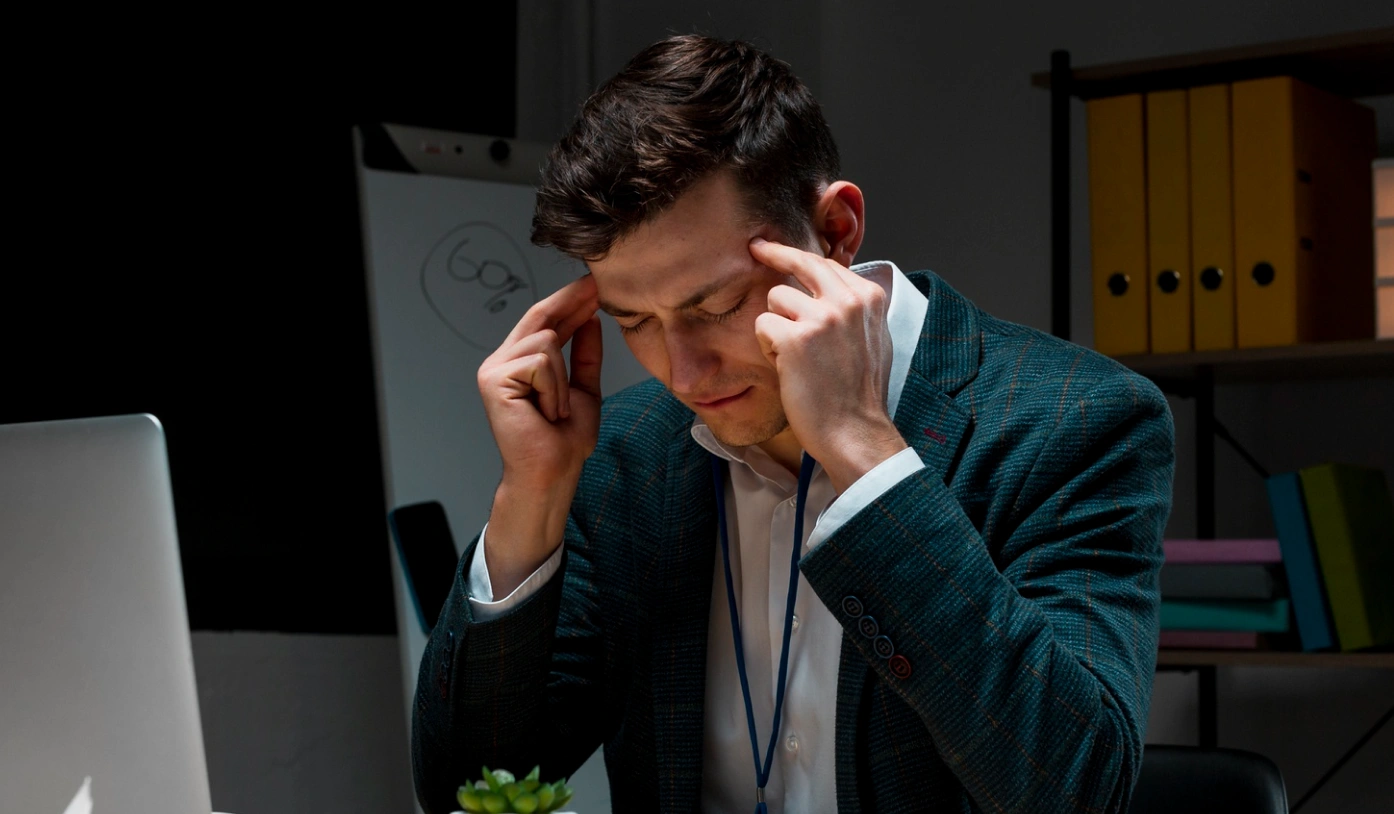 Young man in a suit sitting at a desk, pressing his fingers to his temples with eyes closed, appearing stressed.